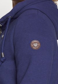Marineblauwe hoodie met een gestructureerde patch met een cirkelvormig ontwerp. Inclusief een koord met geweven details en een metalen punt.