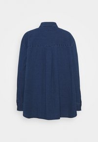 Chemise en denim bleu foncé, manches longues, col pointu, détail de pli dans le dos et fentes sur les côtés au niveau de l'ourlet. Couture simple le long des coutures.