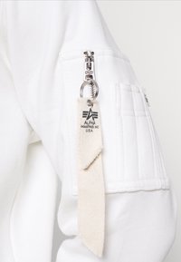 Felpa bianca con una cerniera visibile sulla manica sinistra. Presenta un'etichetta beige con logo e testo stampati, tessuto strutturato e dettagli delle tasche.