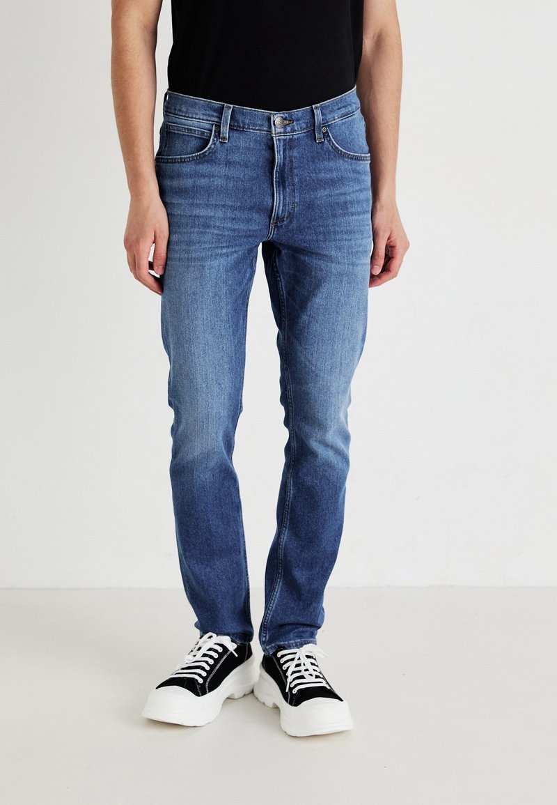 Personne portant un jean slim bleu et des baskets basses noires à semelles blanches épaisses, debout devant un fond clair uni.