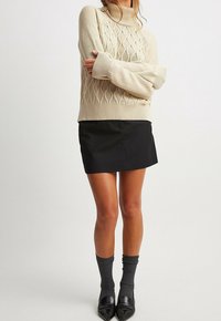 Beżowy sweter z golfem wykonany na drutach, w wzór rombów, w połączeniu z czarną mini spódniczką. Modelka nosi szare skarpetki oraz czarne buty na szpilkach.