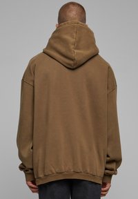 Sudadera marrón de gran tamaño con una capucha grande, mangas largas y dobladillo elástico. Textura suave, con un detalle mínimo, vista desde atrás.