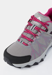 Zapato de senderismo gris con acentos rosas, que cuenta con una parte superior de malla, capuchón de goma en la punta y suela texturizada para mayor agarre. Los cordones son de un rosa brillante con detalles en negro.
