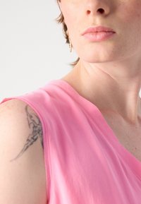 Roze mouwloos topje met een V-hals, gemaakt van zachte stof. Heeft zichtbare stiksels en een tatoeage op de arm. Neutrale achtergrond.