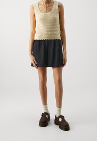 Débardeur beige crocheté à bords festonnés, associé à une jupe bleu foncé. Chaussures compensées marron mises en valeur par des chaussettes claires.