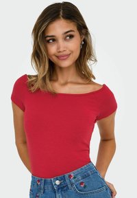 Röd figursydd topp med korta ärmar och vid halsringning, i kombination med blå denimjeans som har blommönster broderade på fickorna.