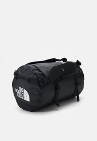 Sac de voyage noir en forme arrondie, fabriqué en matériau durable, avec des sangles ajustables, des boucles et un logo blanc bien visible.