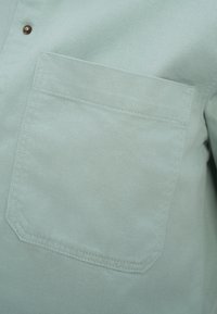 Chemise boutonnée vert clair avec une poche plaquée, un ourlet droit et un tissu finement texturé. Détail de bouton en bronze ajoutant un contraste.