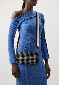 Borsa a tracolla in bianco e nero a fantasia con forma rettangolare, dotata di patta superiore e tracolla a contrasto su un vestito blu a coste.