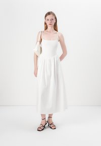 White dress with fitted bodice, smocked texture, and flowing skirt. Model carries a small white bag, wearing black strappy heels.