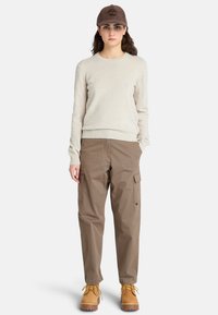 Maglione beige, pantaloni cargo marroni e stivali gialli. Il cappello è marrone con un logo in rilievo. Caratterizza una vestibilità comoda e numerose tasche.