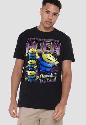 Junger Mann trägt ein schwarzes T-Shirt mit farbenfroher Grafik von vier grünen Aliens mit drei Augen aus "Toy Story" und einem Schriftzug, der "The Claw" referenziert.