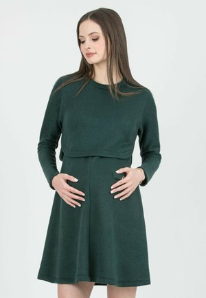 Zwangere vrouw met een donkergroene jurk met lange mouwen, handen rustend op de buik, kijkend naar beneden, staand tegen een effen lichte achtergrond.