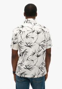 Camisa de manga corta de tela ligera con fondo blanco y patrón de hojas de bambú negras. Presenta un diseño con cuello y botones.
