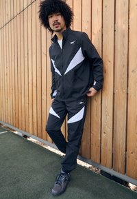 Schwarz-weiße Trainingsjacke mit einem Reißverschluss und einer Hose mit abgewinkelten weißen Akzenten, aus leichtem Material gefertigt. Sneakers sind sichtbar.