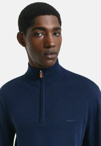 Marineblauwe gebreide sweater met rits, gemaakt van zacht materiaal, met een hoge kraag en subtiele branding op de borst, voorzien van een contrasterende ritstrekk.
