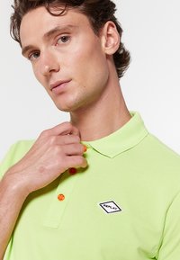 Jonge man die de kraag van een lichtgroen poloshirt met oranje knopen en een wit "Replay" logo op de borst aanpast.