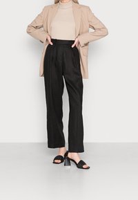 Béžový šitý blazer přes světle béžový top, doplněný černými vysokými širokými kalhotami a černými sandály na podpatku s tkanými pásky.