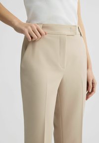 Pantaloni beige su misura con una finitura in tessuto liscia, dotati di una vita a media altezza, tasche frontali e dettagli di cucitura puliti.