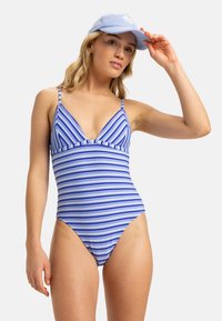 Maillot de bain rayé dans des tons de bleu et blanc, doté de fines bretelles ajustables et d'un dos décolleté. Associé à une casquette bleu clair.
