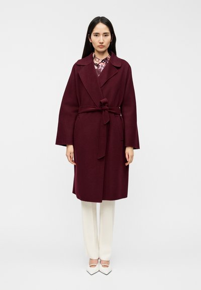 Manteau portefeuille bordeaux avec une ceinture nouée, de larges revers et des manches longues, associé à un pantalon blanc et des chaussures à bout pointu. Texture mate et coupe ajustée.