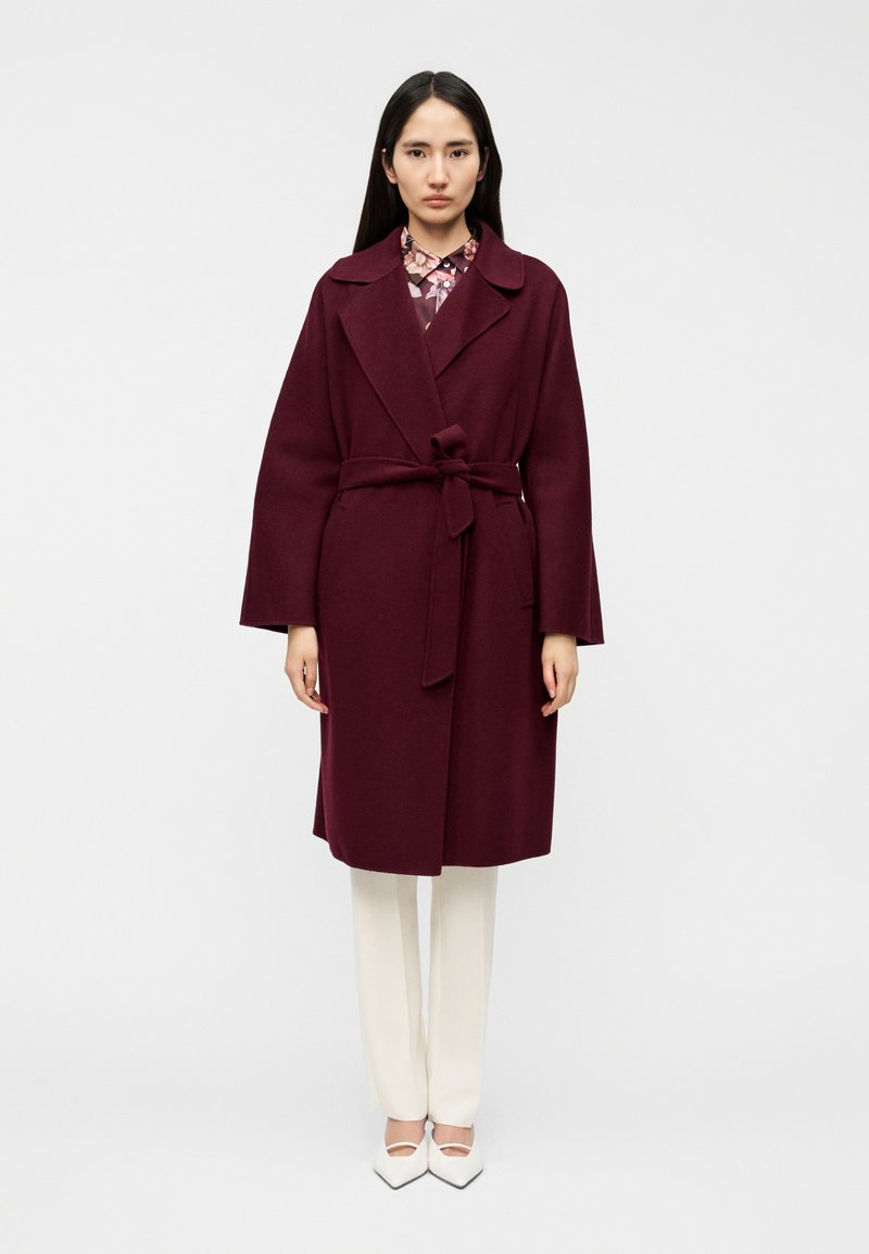 Μπουφάν μπορντό με ζώνη δέσιμο, φαρδιές πέτσες και μακριά μανίκια, σε συνδυασμό με λευκό παντελόνι και παπούτσια με μυτερή μύτη. Λιτή υφή και εφαρμοστή γραμμή.