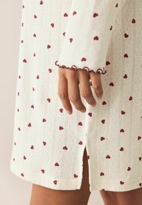 Valkoinen neulemekko, jossa on punaisia sydänkuvioita, ribbitekstuuri ja kynnen muotoiset hihansuut. Mukana on myös sivuhalkio yksityiskohtana.