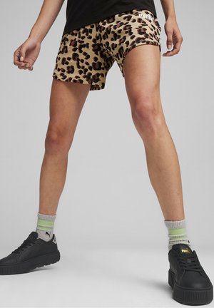Persoon draagt luipaardprint Puma-shorts, zwarte Puma-sneakers en grijze sokken met limoengroene strepen, staand met gespreide benen.