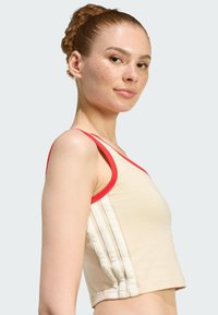 Top corto in tessuto beige con rifiniture rosse. Presenta tre strisce bianche lungo il lato. Tessitura liscia, design aderente.