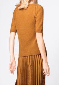 Kvinna iklädd en senapsgul kortärmad stickad topp och matchande plisserad kjol, sedd bakifrån mot en enkel bakgrund.