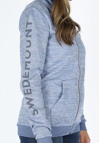 Light blue zip-up sweatshirt with a textured pattern, featuring a high collar, side pockets, and a printed logo along the left sleeve.