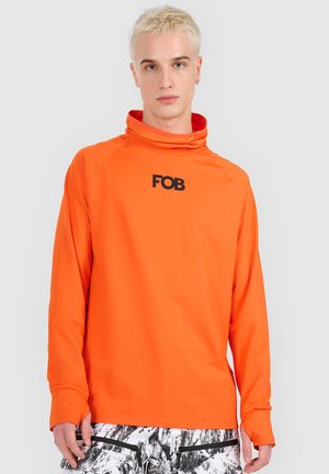 Młoda osoba z krótkimi blond włosami nosi jasną pomarańczową bluzkę z długim rękawem z logo "FOB" oraz czarno-białe spodnie we wzory.