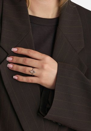 Teemantkujuline hõberõngas peene detailitööga, kantud käel, mille küüned on heledalt roosad, taustal pruunikirju triibuline bleiser.