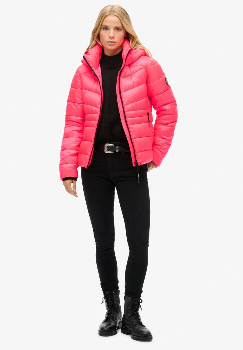 Rosa Puffjacke mit horizontaler Steppung, hohem Kragen und Kapuze, kombiniert mit einem schwarzen Pullover, Jeans und Schnürstiefeln. Glatte Textur.