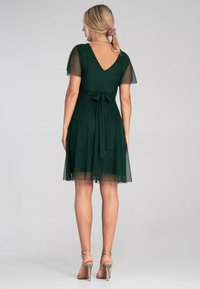 Dunkelgrünes Kleid mit kurzen transparenten Ärmeln, V-Ausschnitt, tailliertem Schnitt und Schleifendetail. Fließender Rock, der bis zur Mitte des Oberschenkels reicht. Kombiniert mit Riemchenschuhen.