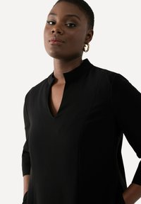Černé šaty z hladkého materiálu mají mandarínkový límec, výstřih ve tvaru V a tříčtvrteční rukávy, čímž zdůrazňují elegantní siluetu.