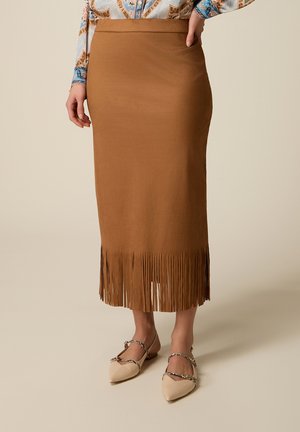 Donna che indossa una gonna midi marrone con orlo a frange e ballerine beige con cinturini intrecciati, in piedi davanti a uno sfondo semplice.