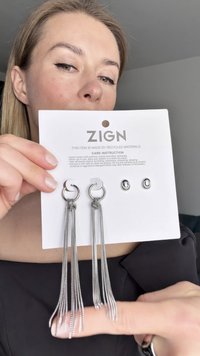 Orecchini a cerchio in argento con catene pendenti, attaccati a un cartoncino. Il cartoncino mostra le istruzioni per la cura e il nome del marchio 'ZIGN'. Realizzati con materiali riciclati.