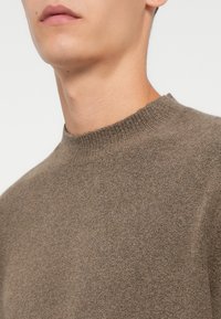 Brązowy sweter dzianinowy z wysokim kołnierzem, teksturowaną powierzchnią i luźnym krojem. Materiał wydaje się miękki i lekko melanzowy.