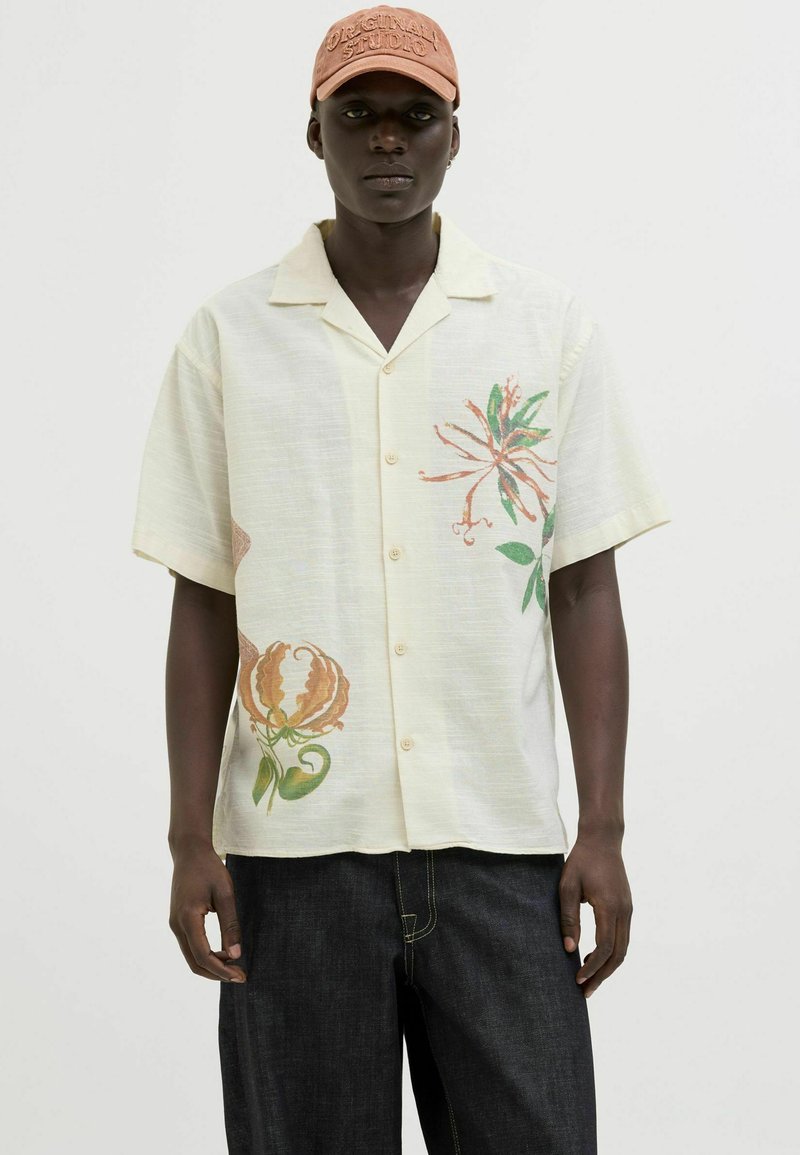 Camisa de manga corta, de color amarillo claro con patrones florales en naranja y verde; con botones al frente, corte relajado, usada con pantalones de mezclilla oscuros.