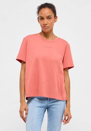 Woman wearing a loose coral T-shirt with faint "Mustang Jeans" text and light blue jeans, standing against a plain light background.