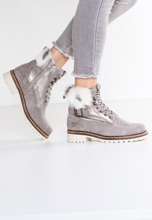 Vrouw draagt grijze suède enkellaarsjes met bontafwerking, ritssluiting aan de zijkant, veters en gesp, gecombineerd met lichtgrijze jeans met rafelige zomen.