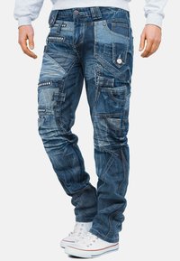 Pantaloni cargo in denim blu con molte tasche, cuciture a contrasto e dettagli decorativi; indossati con sneaker bianche.