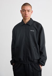 Black zip-up jacket with a collared neck, elastic waistband, two side pockets, and white logo accent. Made of smooth, lightweight material.