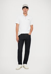 Ung mand iført hvid polo, mørke jeans, hvide sneakers og beige kasket, der står foran en ensfarvet hvid baggrund.