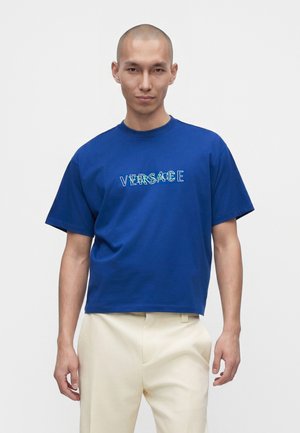 Mand med barberet hoved iført en klarblå Versace T-shirt og lyse beige bukser mod en ensfarvet hvid baggrund.