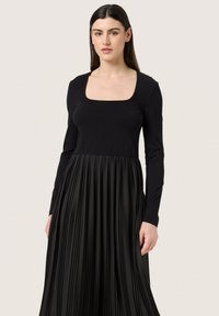 Long-sleeve black dress with a square neckline, featuring a fitted bodice and a pleated skirt. Smooth fabric with a structured design.