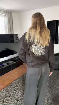 Zwarte sweatshirt met een rond logo op de achterkant, gecombineerd met losvallende grijze broek. Gestructureerde stof en ontspannen silhouet.