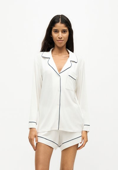Femme aux longs cheveux foncés portant un haut de pyjama blanc boutonné et un short assorti avec un liseré noir, debout devant un fond blanc.