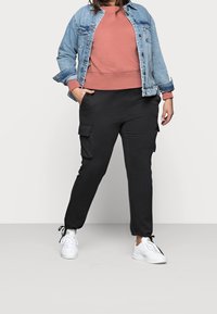 Chaqueta vaquera sobre un sudadera color óxido, combinada con pantalones cargo negros con bolsillos y zapatillas blancas. Un look casual con tejidos texturizados.